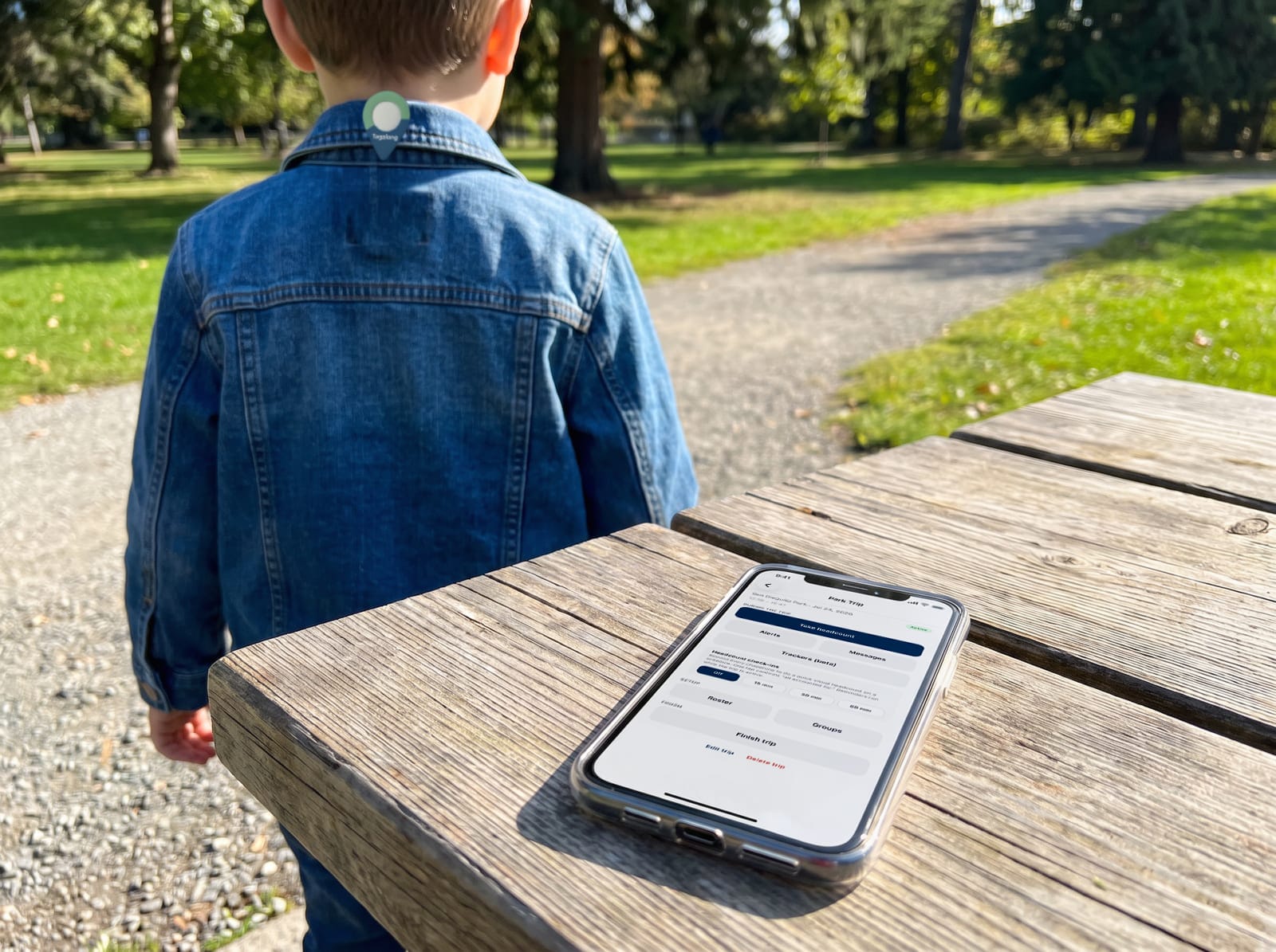 A child wearing a Tagalong tag on their jacket collar, walking ahead of a chaperone's phone resting on a picnic table